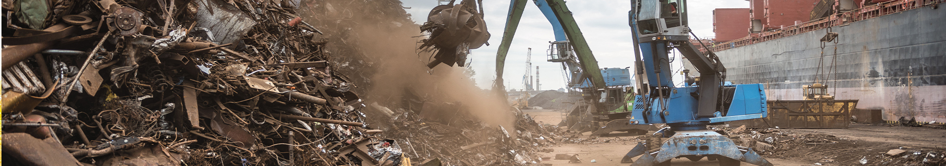 Material handlers equipped with waste and recycling tires sorting and moving scrap metal at a recycling facility near a docked ship.